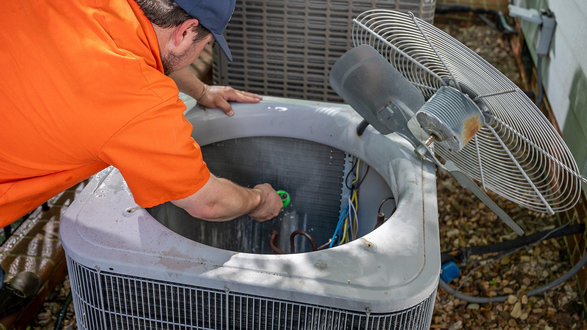 HVAC contractor in orange shirt repairs outdoor residential HVAC unit. 